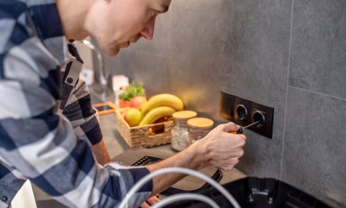 Photo en gros plan d'un homme qui touche une prise noire sur le mur de la cuisine avec un tournevis spécial.